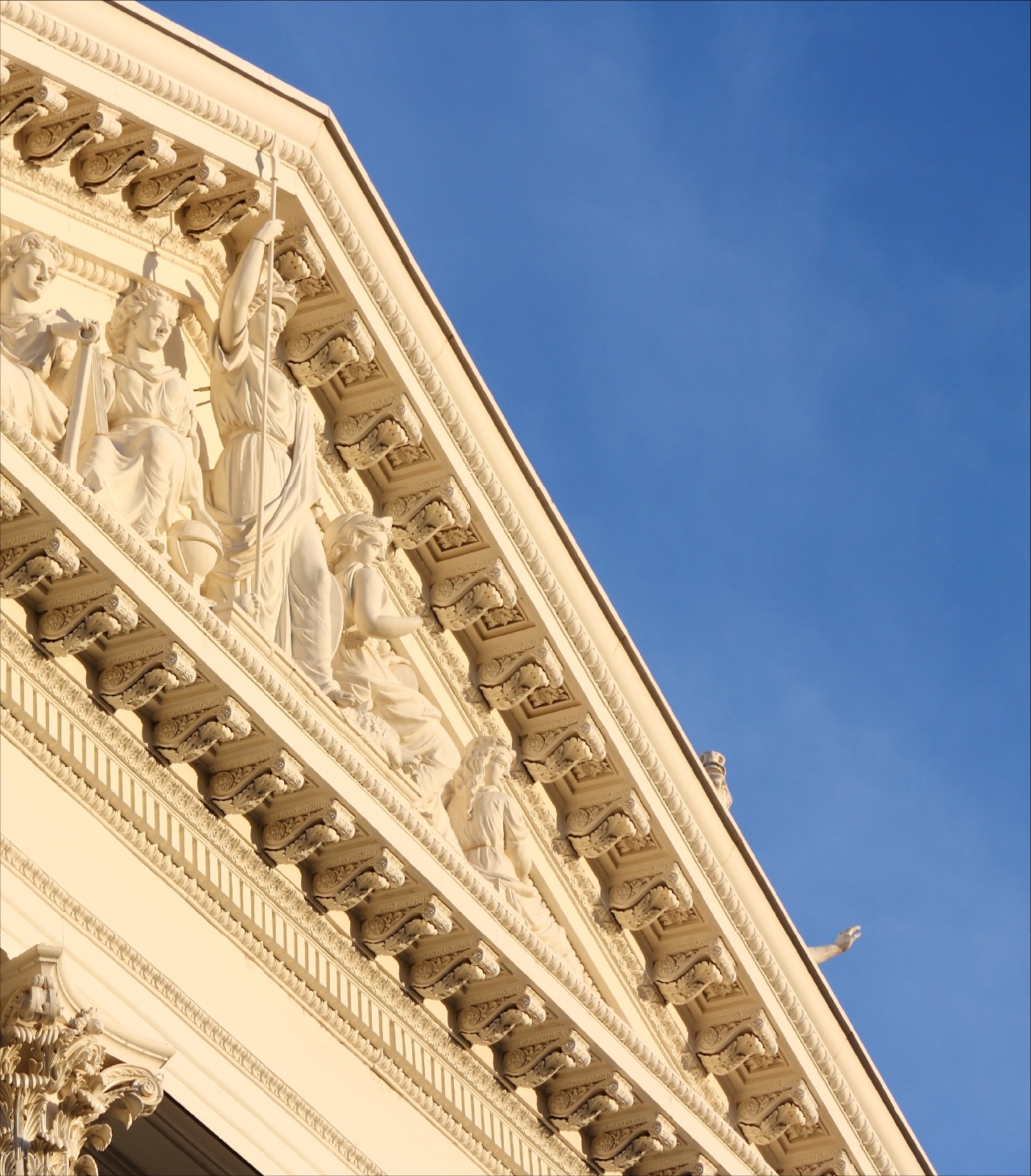 Classical building façade with sculpted figures along a triangular pediment, viewed at an angle against a clear blue sky.